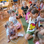 Dzieci siedzą na dywanie podczas akcji Narodowego Czytania, jedna dziewczynka zgłasza się do odpowiedzi.