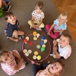 Dzieci siedzą wkoło hula-hop z kolorowymi piłkami, kropkami i kubeczkami.