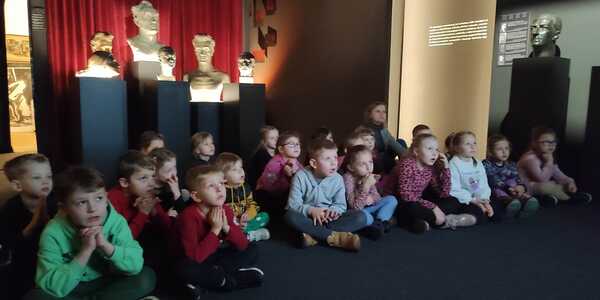 Dzieci oglądają film edukacyjny, w tle popiersia.