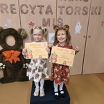 Dwie dziewczynki w misiowych sukienkach stoją na scenie z dyplomami.