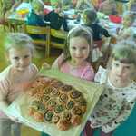 Trzy dziewczynki trzymają blachę z ciastem drożdżowym w kształcie baranka, reszta dzieci patrzy zza stołu.